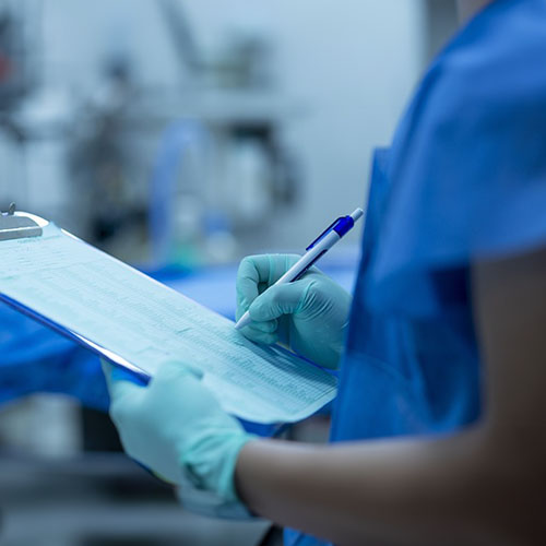 Eine Person im blauen Kitel und Handschuehn steht in einem OP und hat ein Klemmbrett mit einem Formular in der Hand und einem Stift zum schreiben in der anderen.jpg